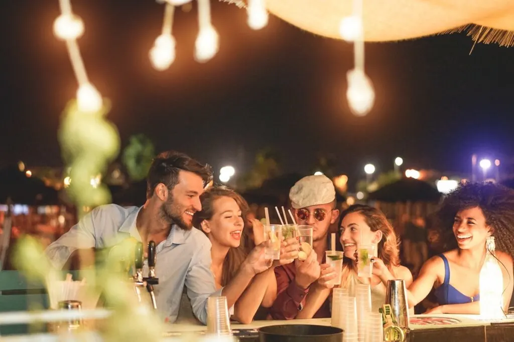 Happy friends cheering and drinking cocktails at beach party outdoor - Young millennials people having fun at weekend summer night - Youth lifestyle and nightlife concept - Main focus on left guys