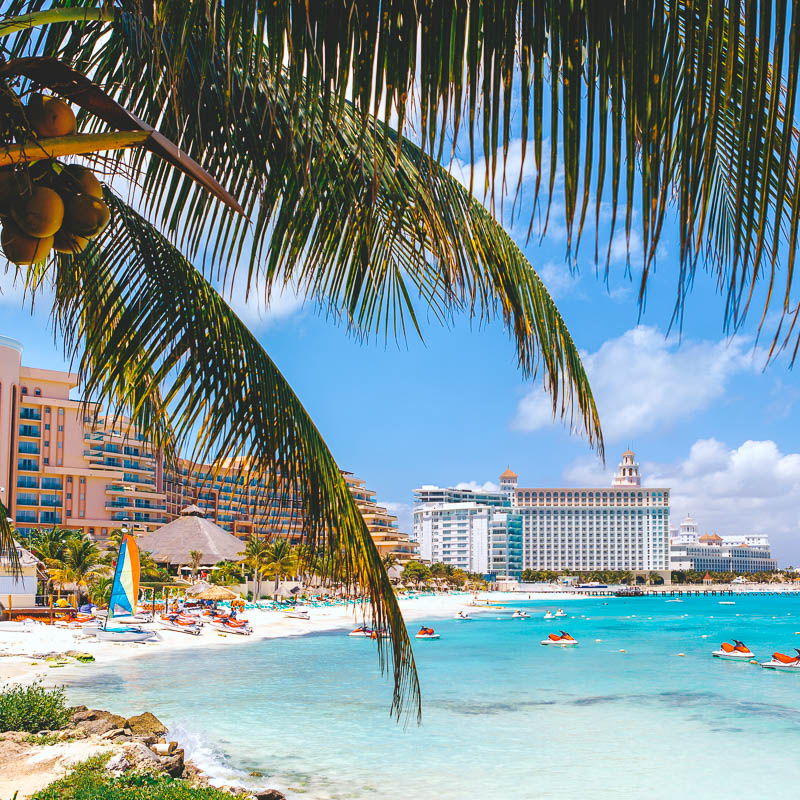 Cancun beach with turquoise blue water and jet skis in the water and hotels in the background.