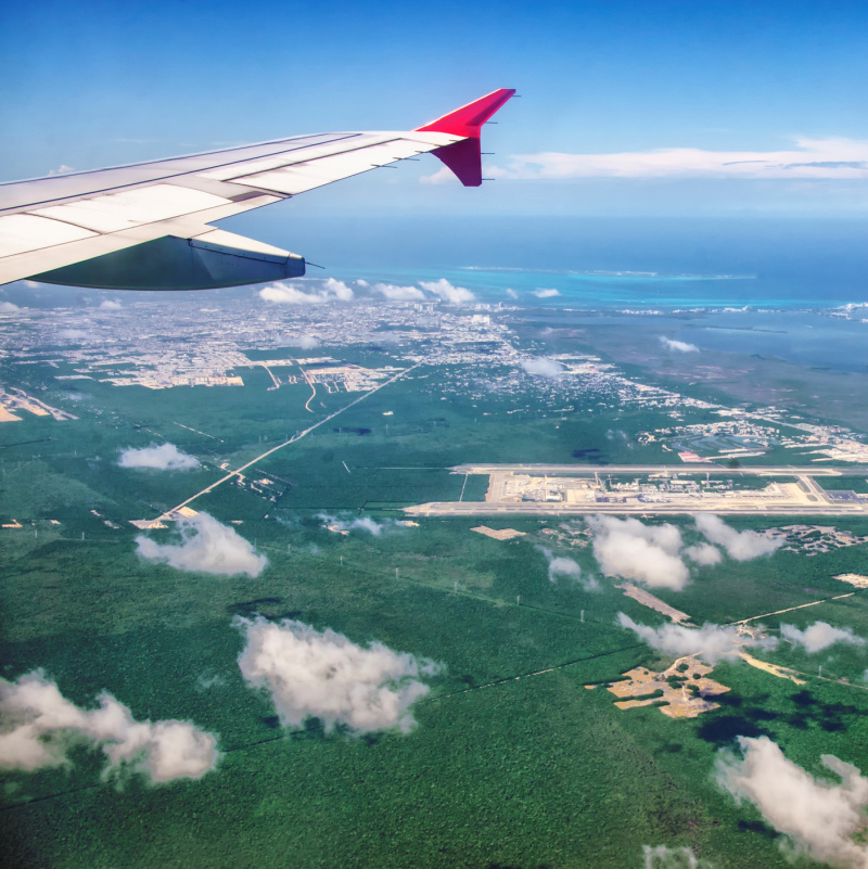 view of Cancun during the day from window seat of airplane, clouds in the foreground and grass and beach can be seen in the distance.