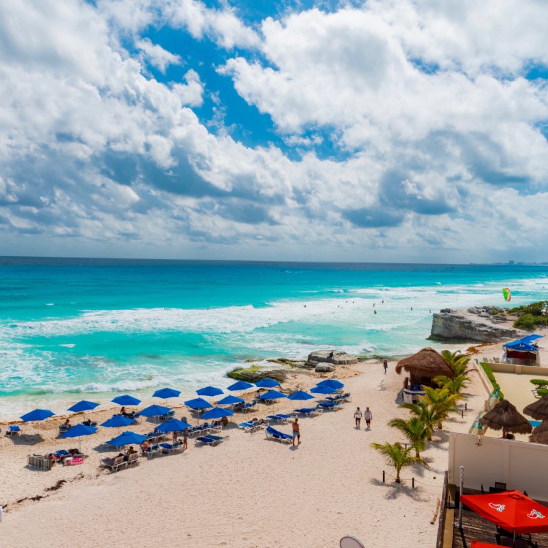 Beach in Cancun, Mexico during the day. Lots of blue beach umbrellas and tourists enjoying the beach.