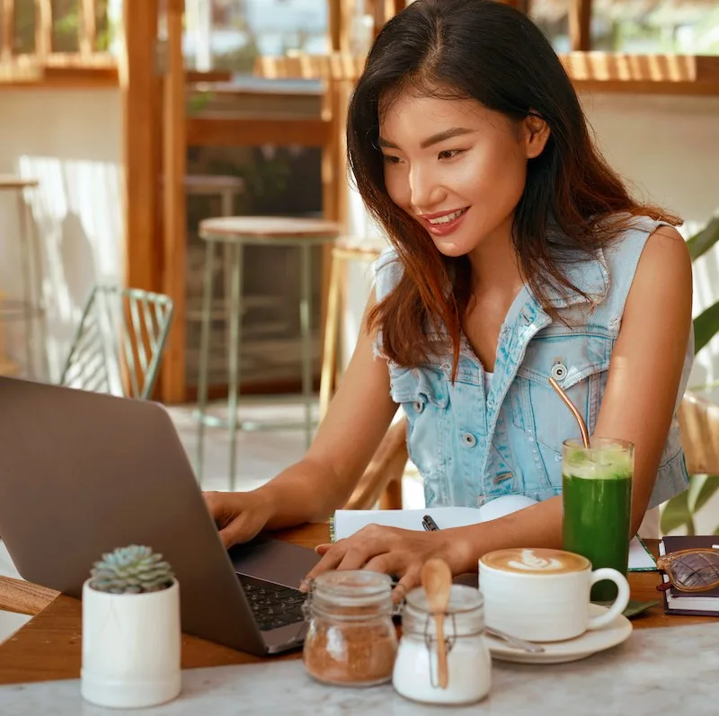 Woman wearing sleeveless jean shirt working on her laptop at a sunny cafe. digital nomad on vacation.