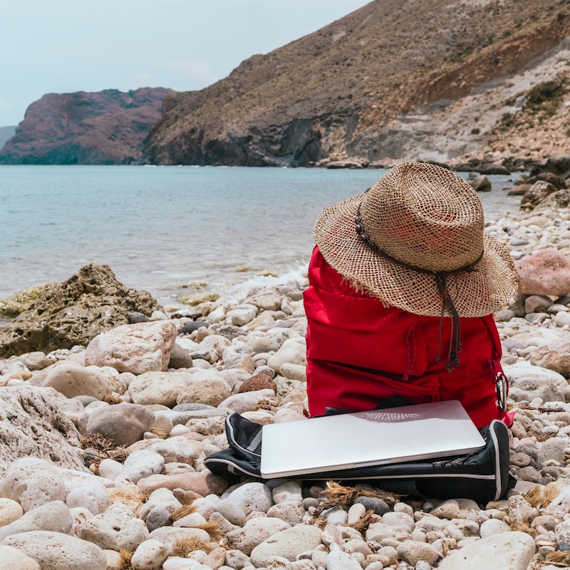 a red adventure and travel backpack with a straw beach hat and a laptop in front of the mediterranean sea. vacation concept and destinations for tourism and teleworking in digital nomads.