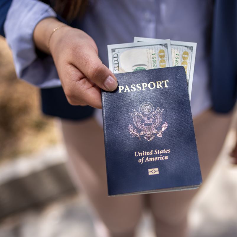 A traveling woman's hand holding a blue American passport with some money inside while wearing business clothes outside on a sunny day