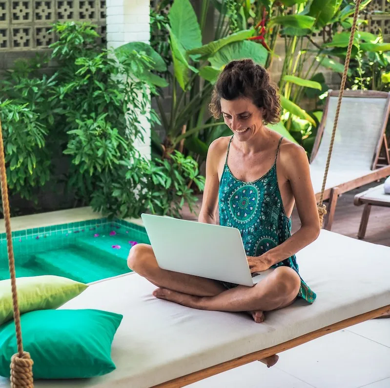 happy working hippie digital nomad woman sitting with laptop on a bamboo hanging swing surrounded by green plants and pool