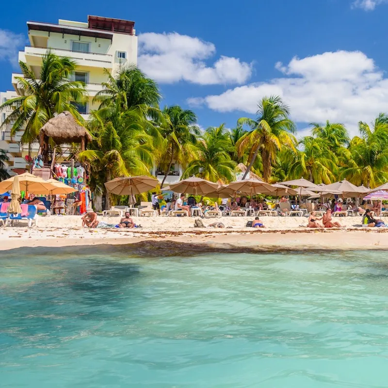 People sunbathing on the white sand beach with umbrellas, bungalow bar and cocos palms, turquoise caribbean sea, Isla Mujeres island