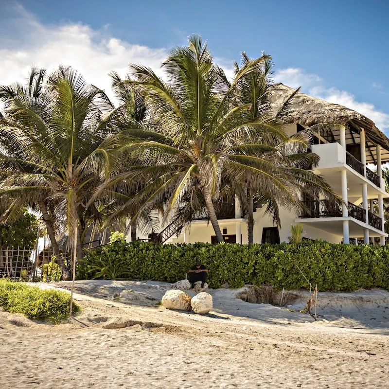 Tulum beach resort surrounded by palm trees with a man sitting in the background on his phone.