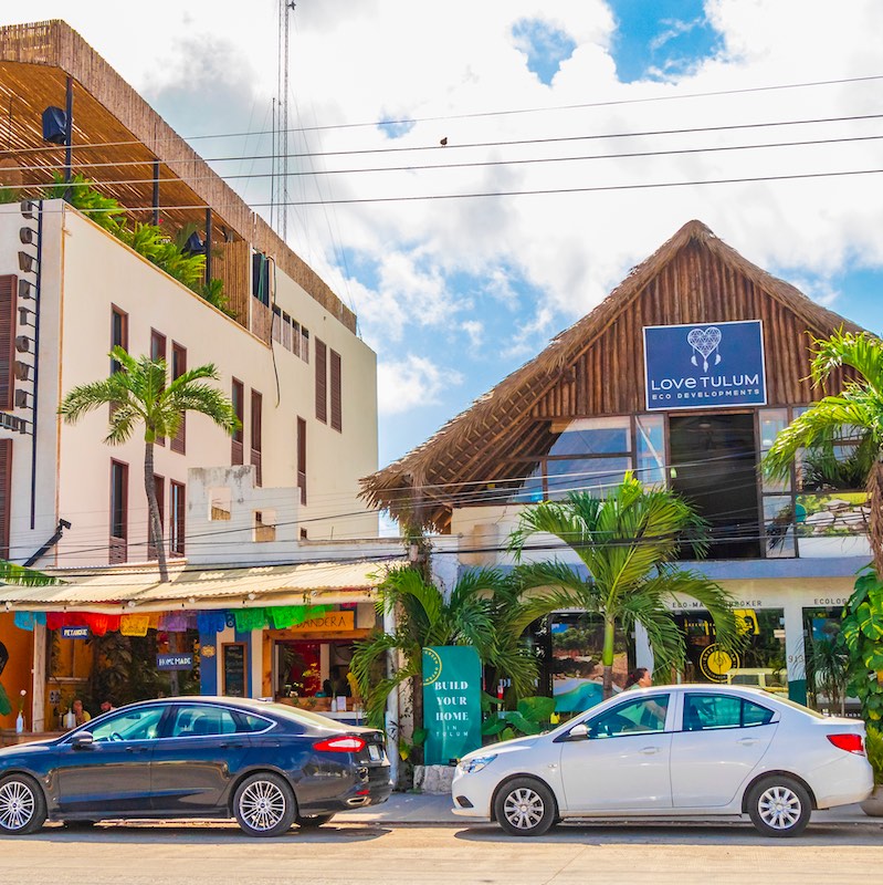 Driving thru typical colorful street road and cityscape with cars traffic palm trees bars and restaurants of Tulum in Mexico.