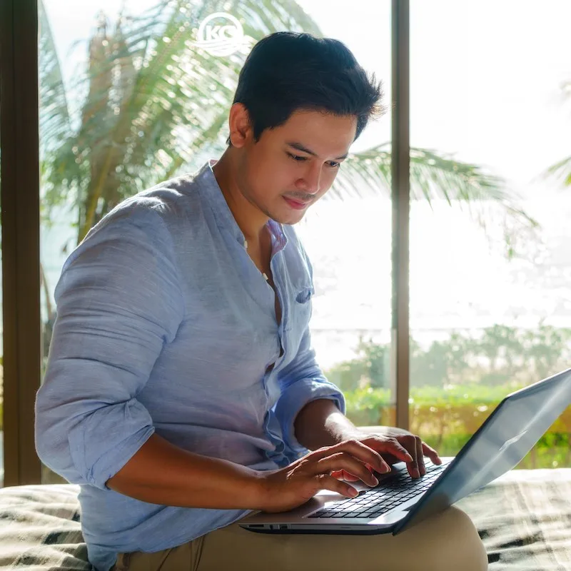 Young Asian businessman sitting in bed and working with laptop in room at resort near sea during a summer vacation holiday travel.