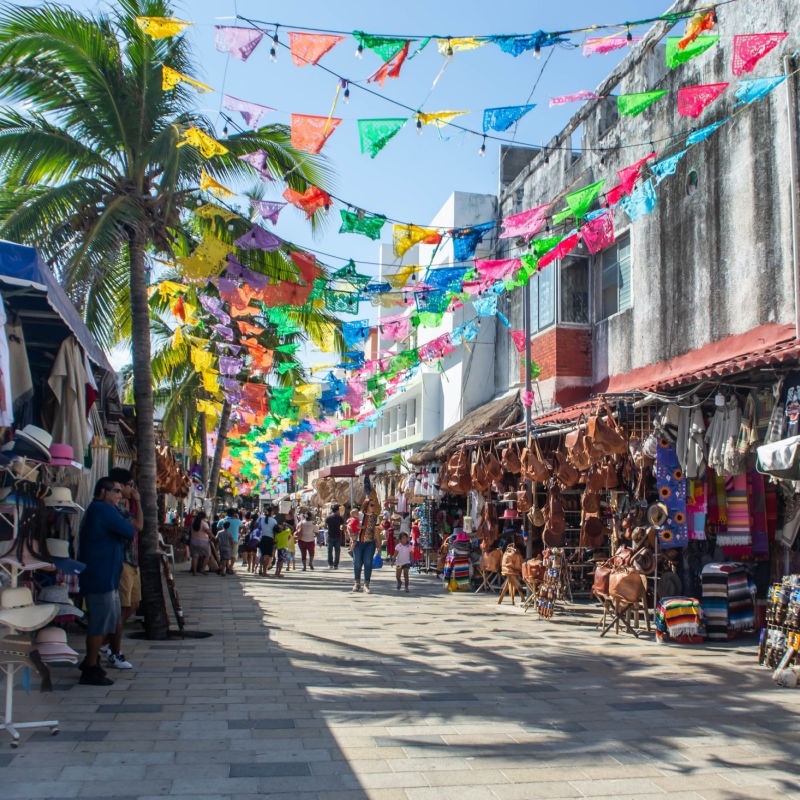 Tourists walking along 5th Avenue in Playa del Carmen, past souvenir shops and other vendors.