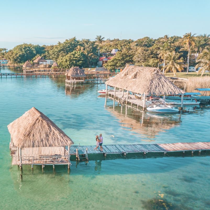 Aerial view of a Couple in Bacalar pier, Riviera Maya, Mexico on clear blue waters by tiki huts and palm trees on sunny day
