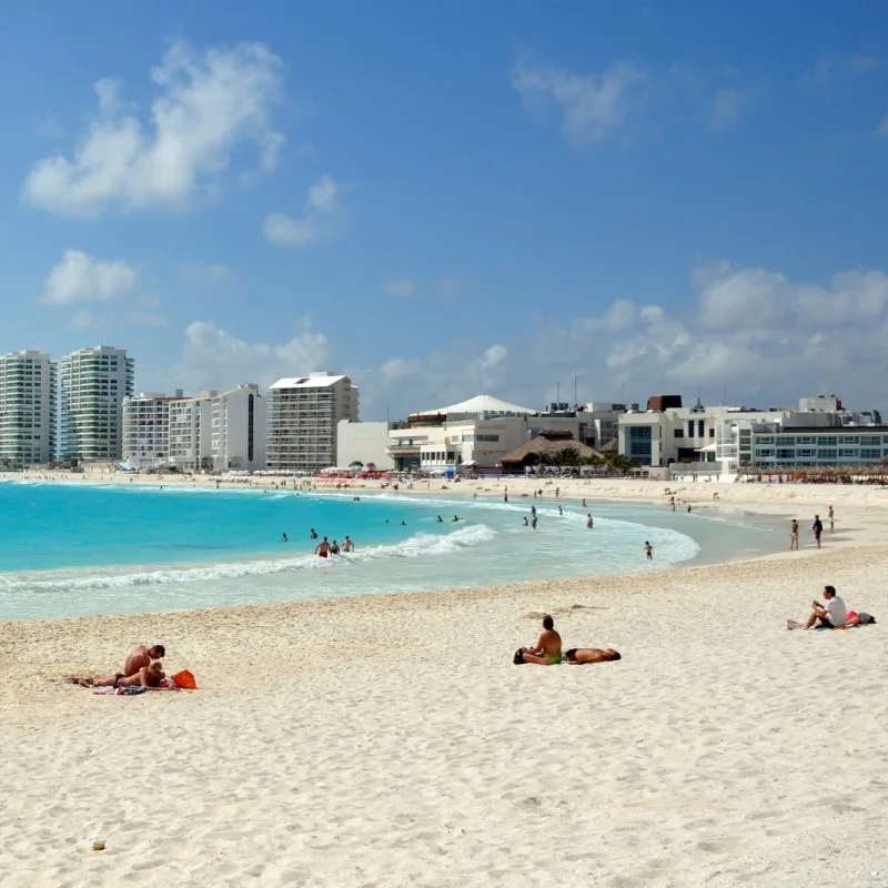 Busy Cancun Beach with people laying on the sand and resorts in the background with a view of aqua blue waters.