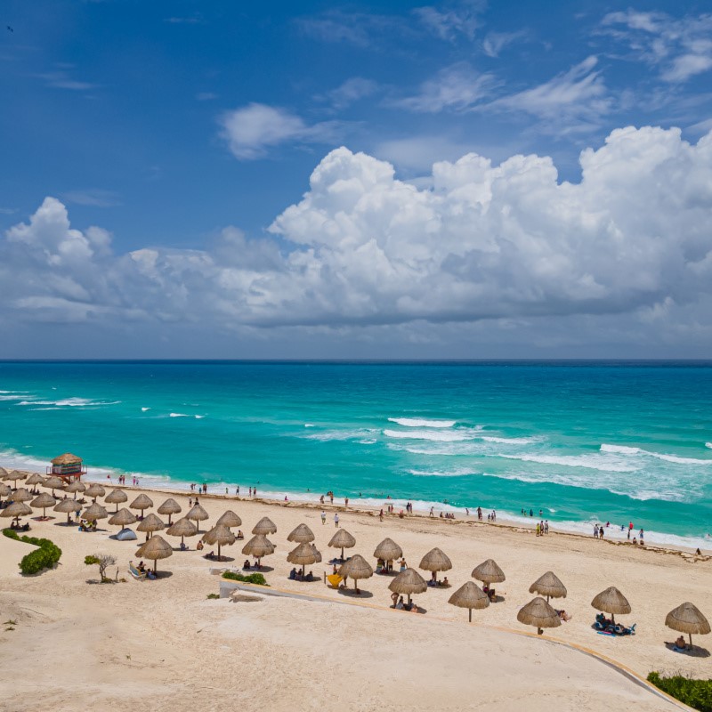 Cancun Beach with people laying in the sand under umbrellas and turquoise blue water in the background.