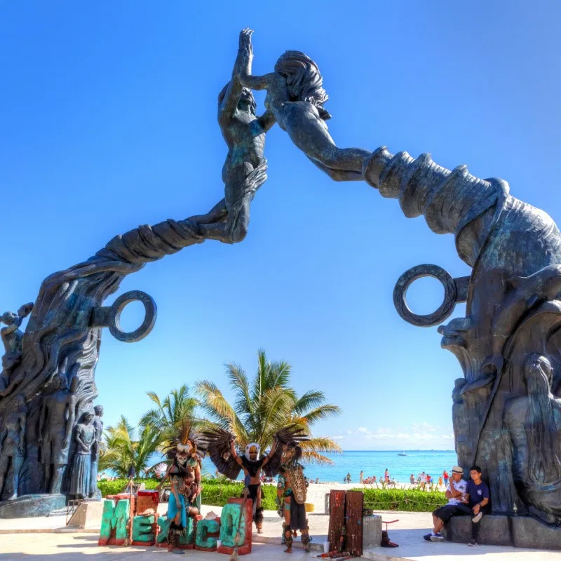 Playa del Carmen Statue with performers and tourists standing in front and the beach in the background.