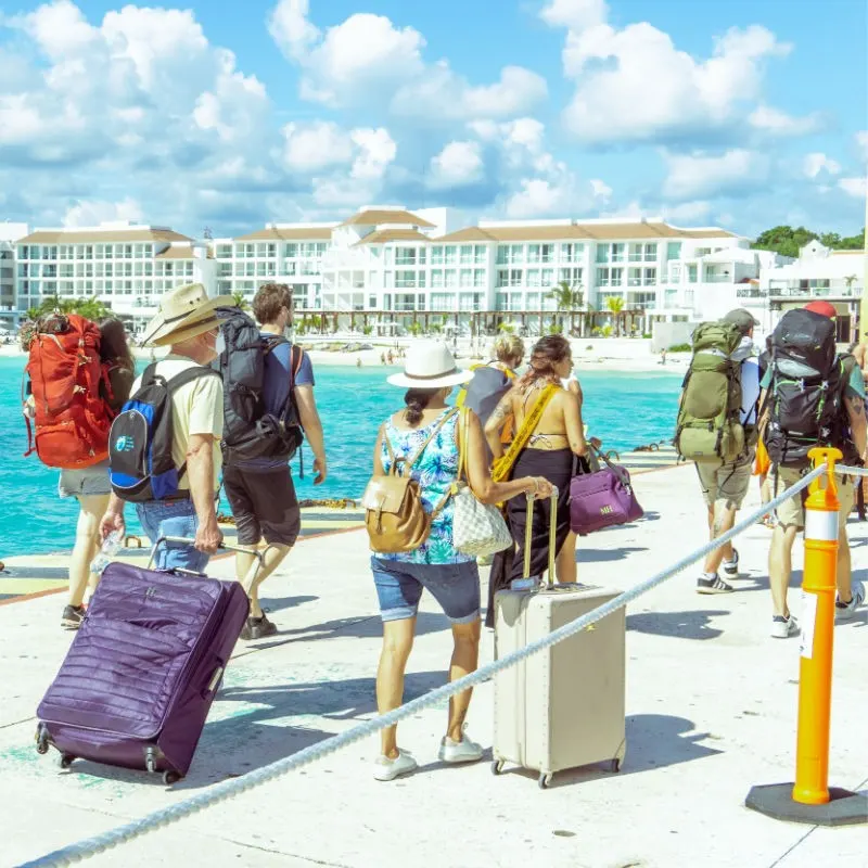 Tourists with Luggage walking by the water on a bridge in Playa del Carmen, with a resort in the background.