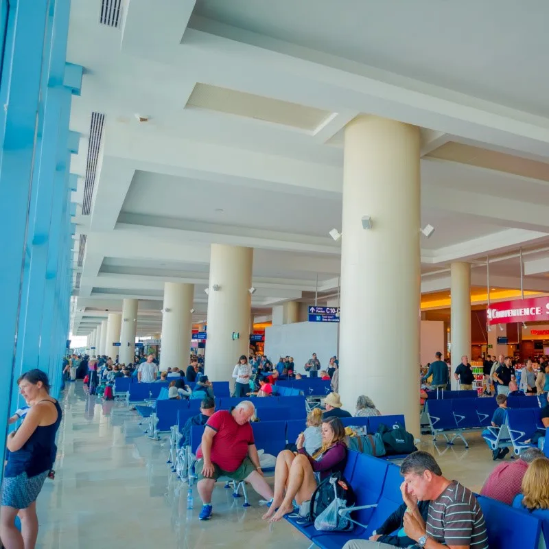 Travelers Waiting for Flights at Cancun Airport. One woman is in the background looking out the window, while others are sitting and waiting.