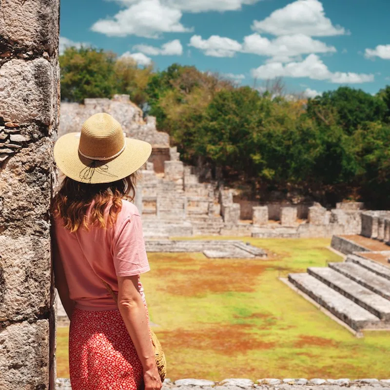 view of tourist wearing dress and hat leaning against a wall looking out at ancient Mayan ruins site in Mexico, sunny day.