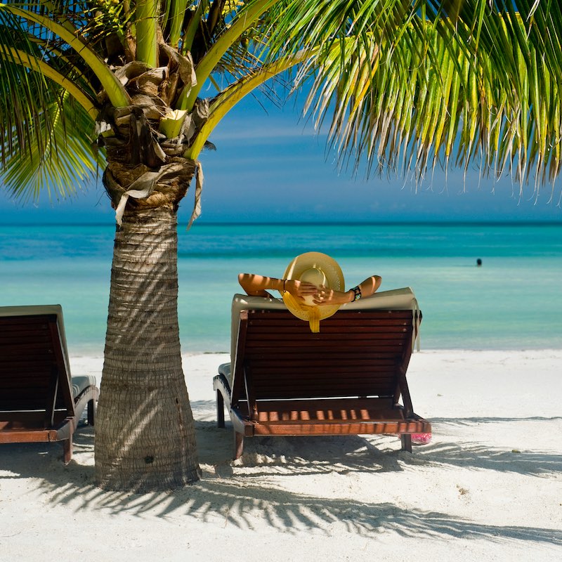 A view from behind of a tourist relaxing on Holbox Island, Mexico, with his hands above his head on a beach lounger under palm trees on the beach, with ocean views in the background.
