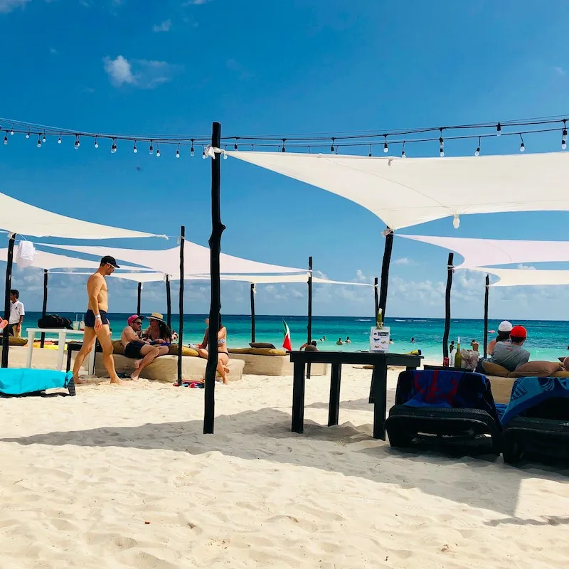 tourists at a private beach club on the beach equipped with straw beds and umbrellas in the Caribbean sea in Mexico with palm trees and white sand.