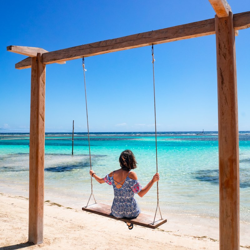 Brunette girl on a swing looking to the horizon, surrounded by a paradise beach with white sand and turquoise water, at Mahahual village, in Quintana Roo, Mexico.