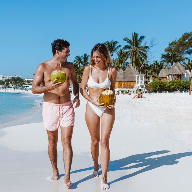 A happy couple walking on a beach drinking from coconuts
