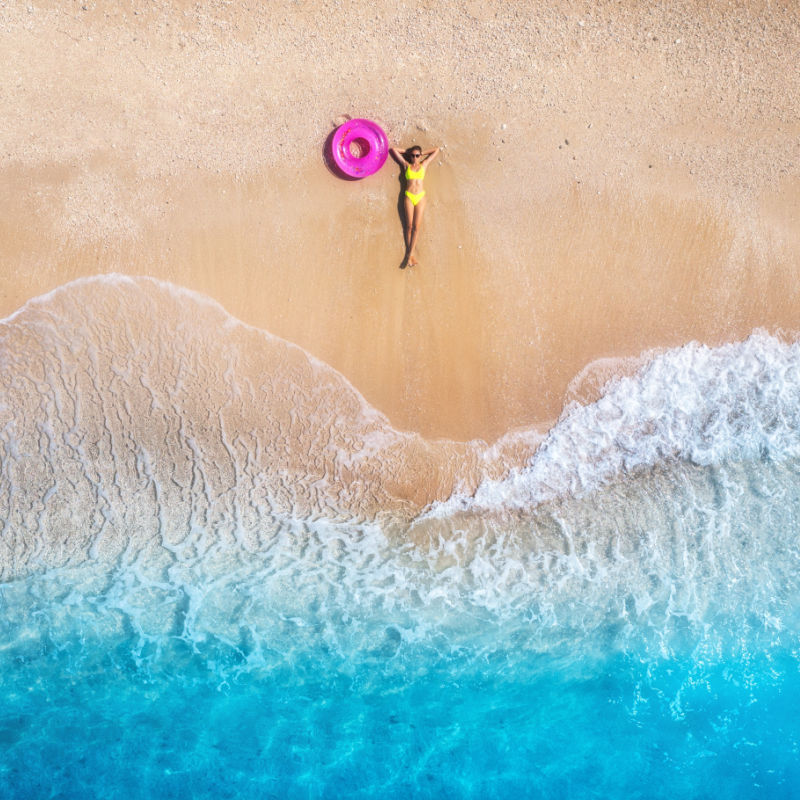 Aerial view of the lying beautiful young woman with pink swim ring on the sandy beach near sea with waves at sunset.