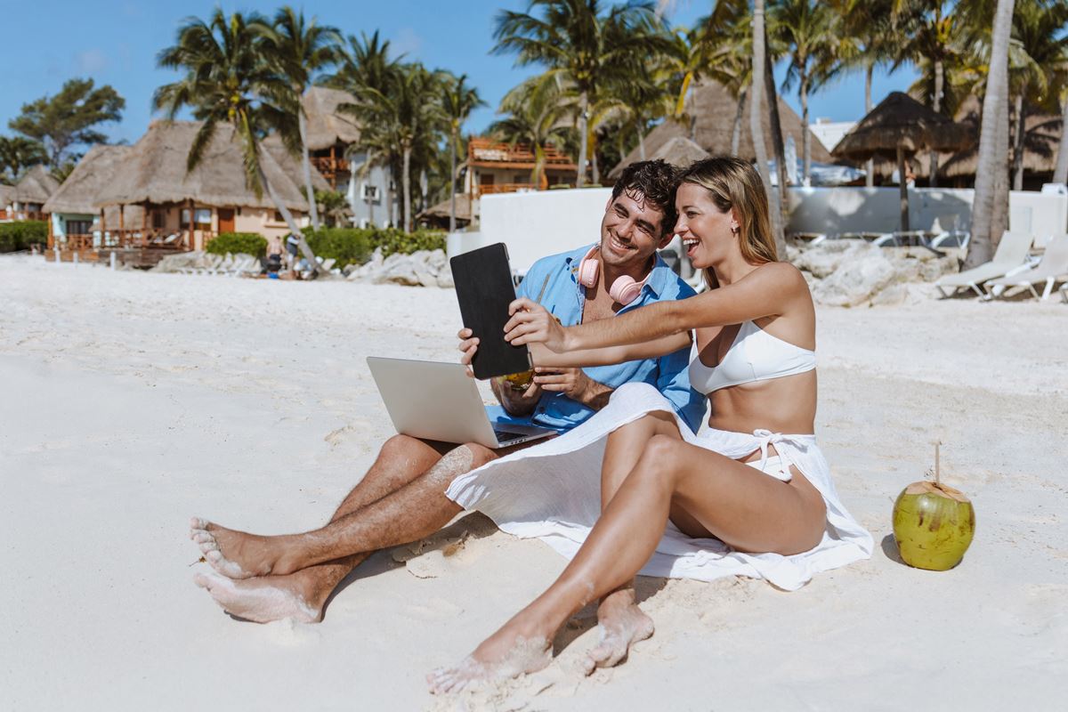 Couple at the beach with laptop and tablet with palapas and palm trees in the background