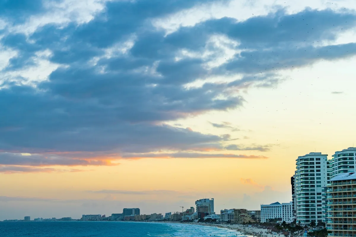 A sunset casts a warm glow over Cancun's beachfront, with scattered clouds reflecting the fading light above the vibrant, bustling shore.