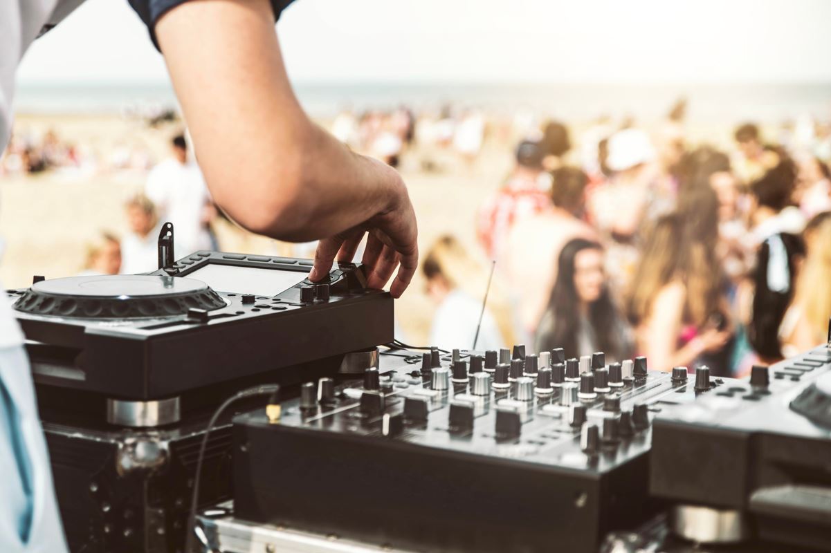 A dj mixing on a set of decks with a shot of people partying on a beach in the background