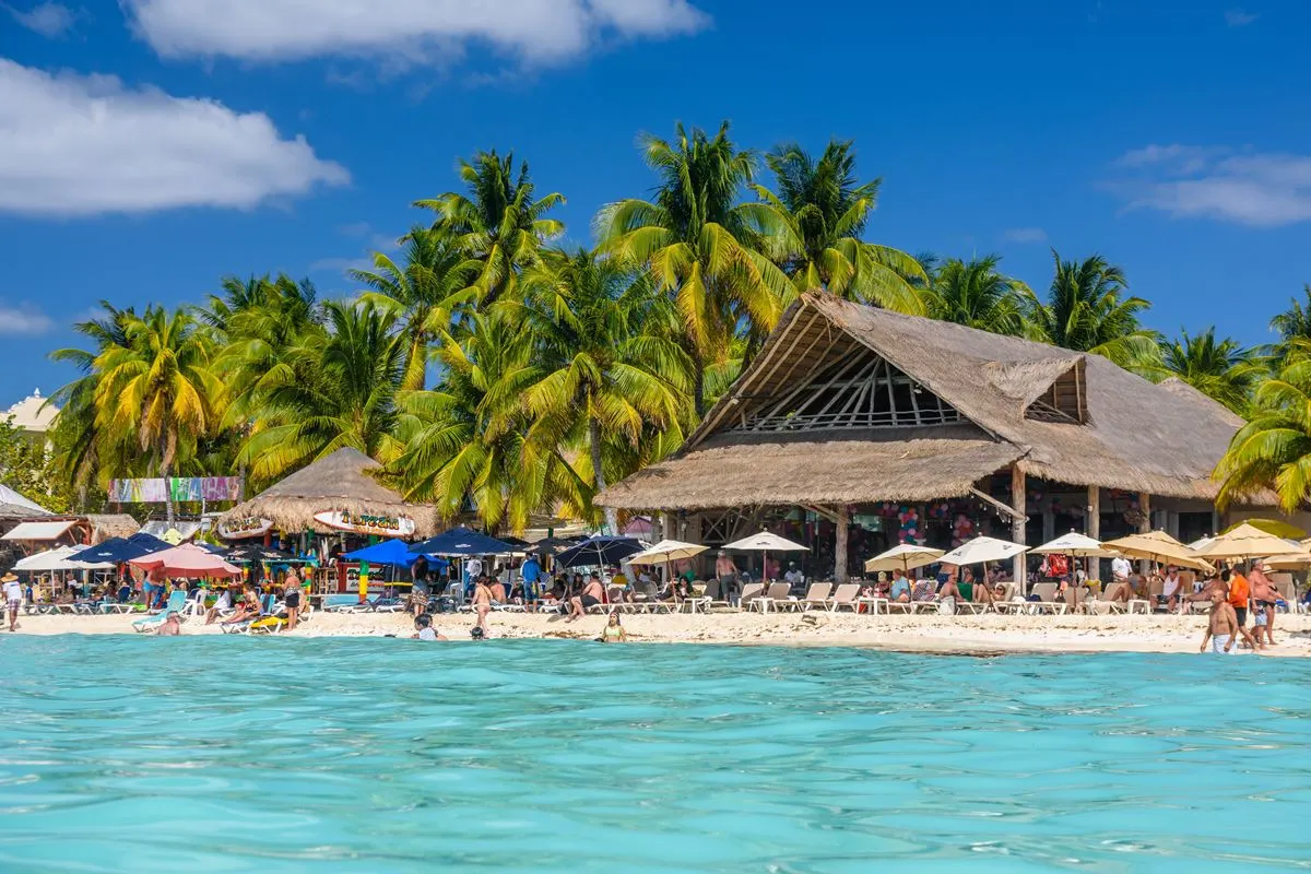 Tourists enyoing a beach day at Isla Mujeres, with turquoise waters and palm trees and a palapa in background