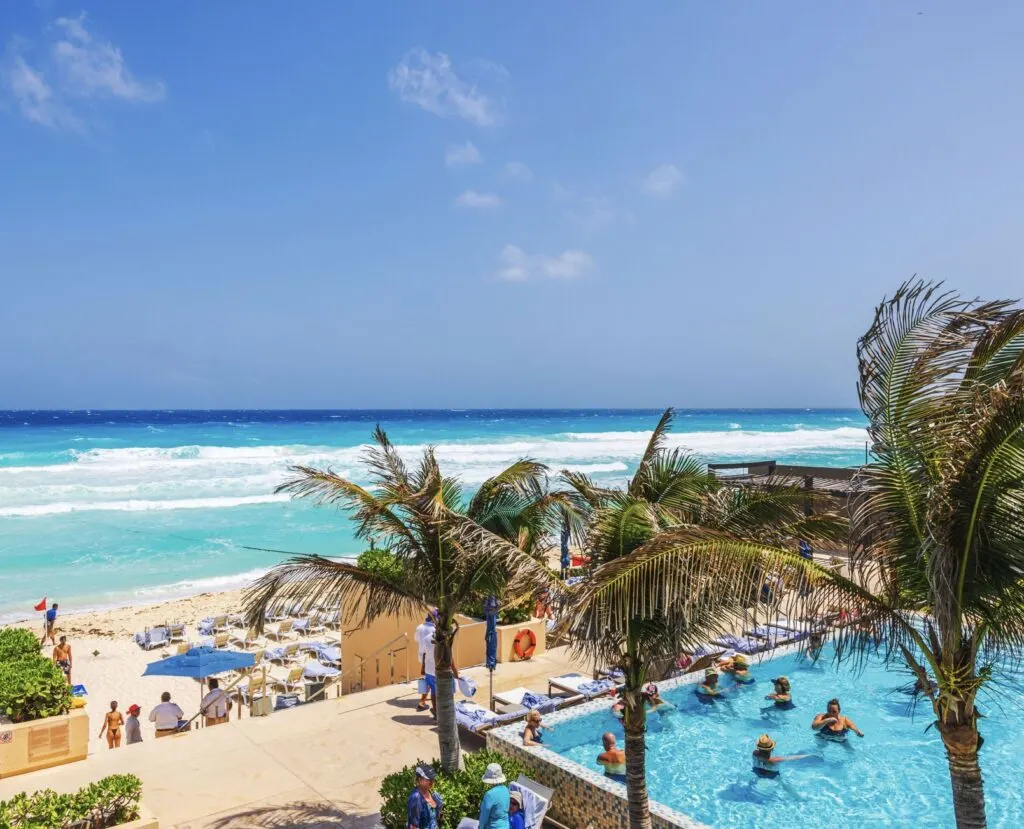 Palm trees swaying near resort pool overlooking turquoise waves of Caribbean Sea with sun loungers and beachgoers. Mexico. Cancun.