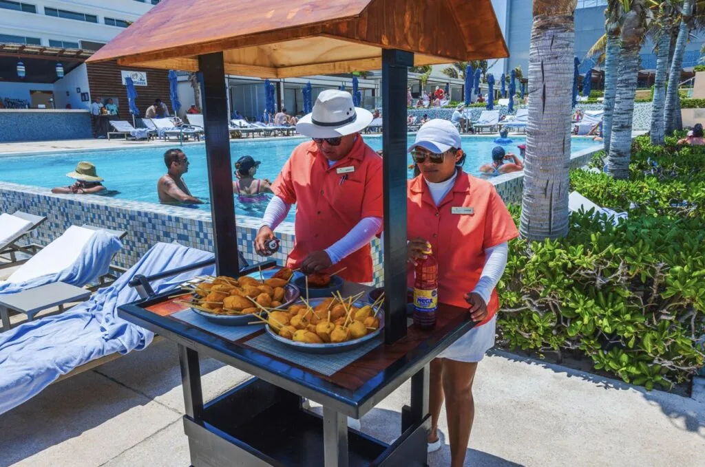 Resort staff preparing peeled mango on skewers at outdoor food cart near pool with lounge chairs and vacationers Cancun. Mexico.