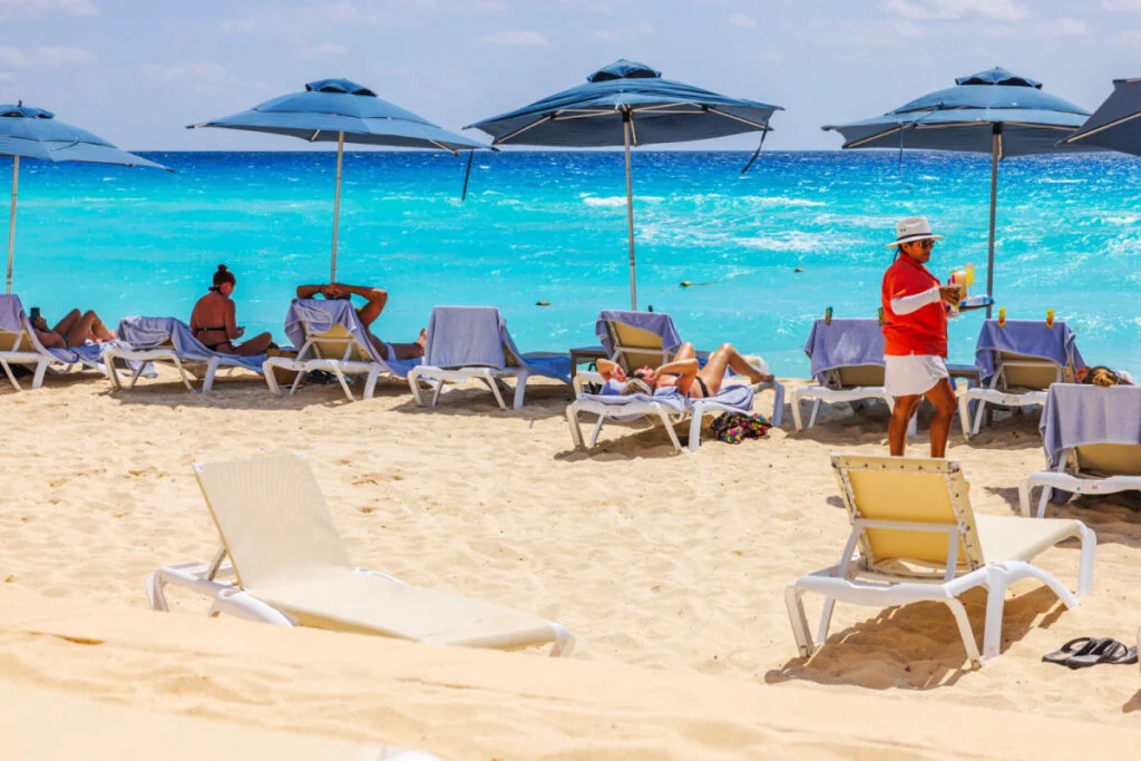 Caribbean beach with tourists relaxing on sun loungers under blue umbrellas while waiter in serves drinks against turquoise sea waves. Mexico. Cancun