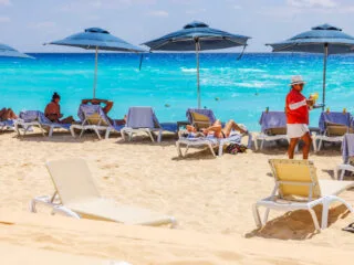Caribbean beach with tourists relaxing on sun loungers under blue umbrellas while waiter in serves drinks against turquoise sea waves. Mexico. Cancun
