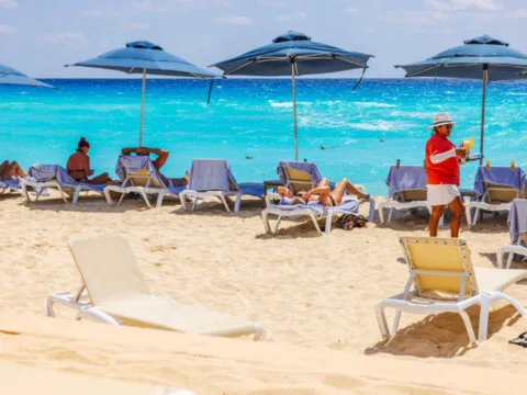 Caribbean beach with tourists relaxing on sun loungers under blue umbrellas while waiter in serves drinks against turquoise sea waves. Mexico. Cancun