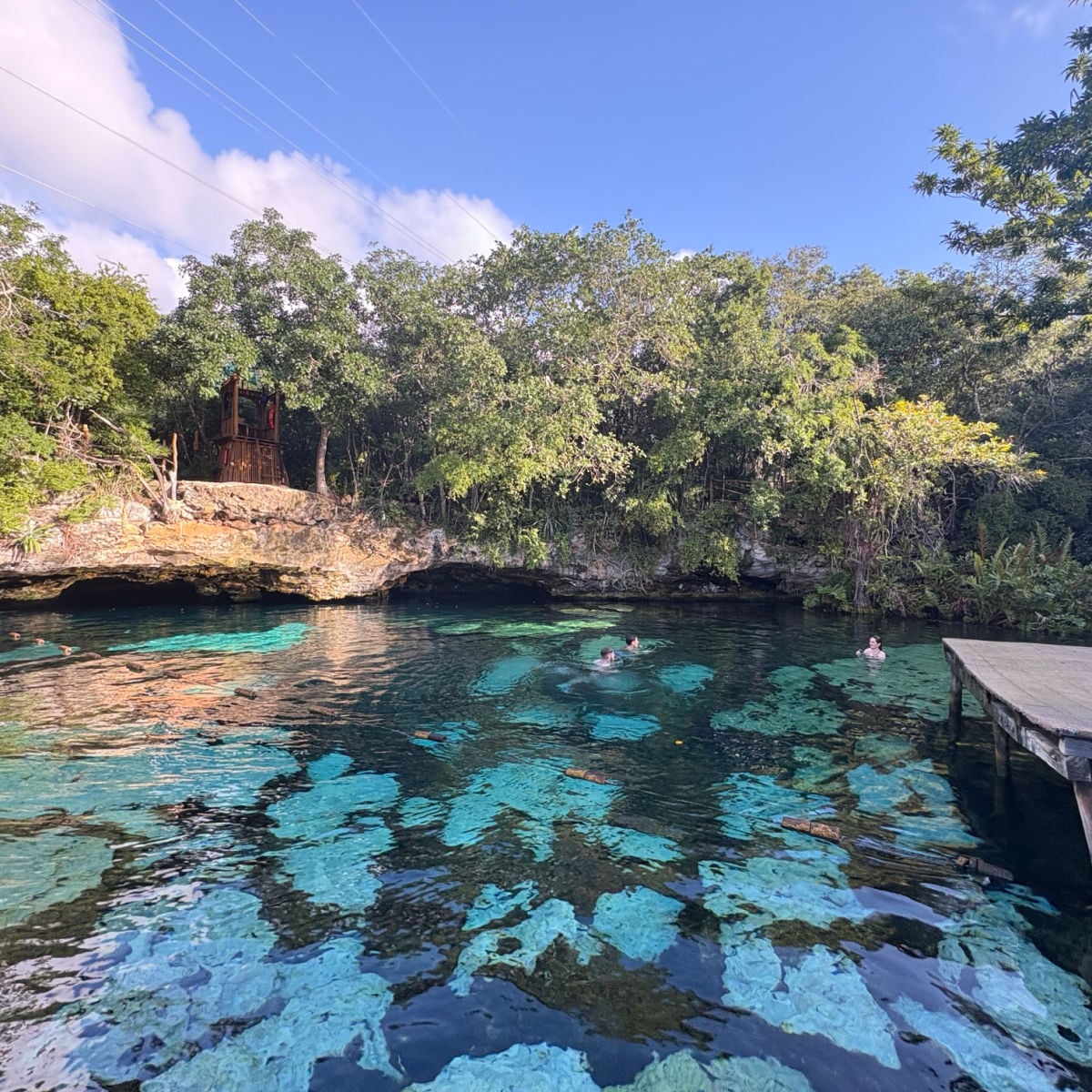 Cenote Azul, a natural freshwater cenote surrounded by lush vegetation, seen in the Riviera Maya. Visitors swim and relax in the clear turquoise water.